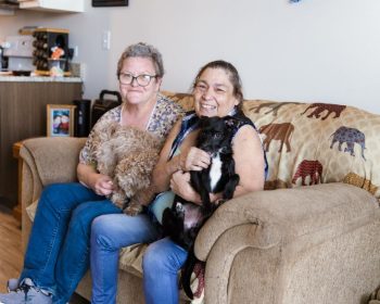 two happy residents of walnut flats, sitting on a couch with dogs in their laps