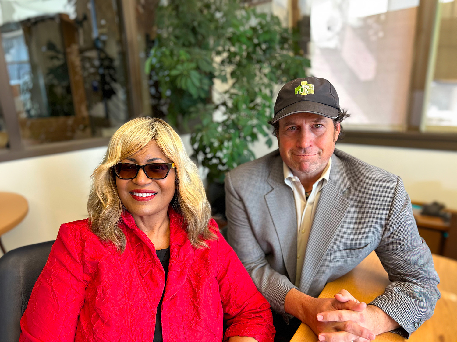 Woman in a red business jacket and sunglasses and a man in a sport coat with a baseball hat sit in a conference room smiling